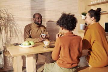 Fototapeta na wymiar Focus on young man with cup of coffee looking at his wife during chat while sitting by wooden table with family in country house