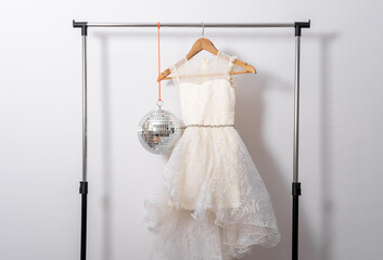 White puffy dress hangs on hanger on clothes rail with disco ball on white wall background.