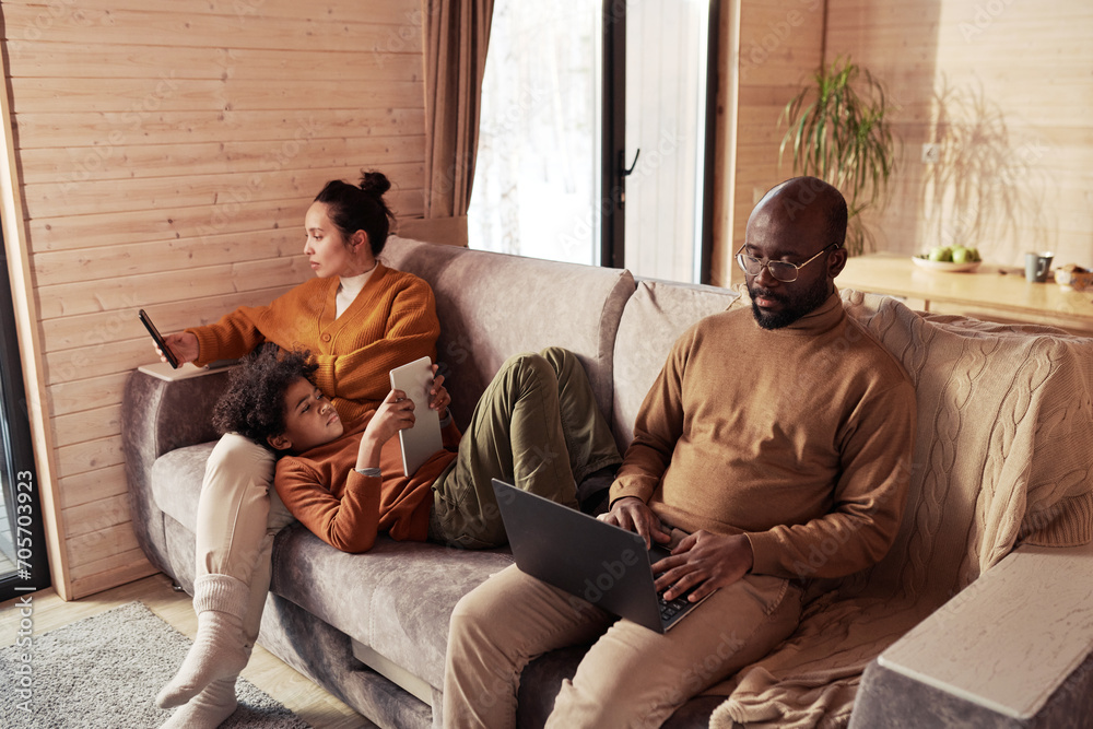 Poster cute boy with tablet relaxing on couch between his mother using mobile phone and father typing on la