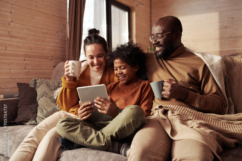 Poster happy young intercultural family of three looking at screen of tablet held by cute boy while sitting
