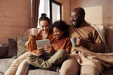 Happy young intercultural family of three looking at screen of tablet held by cute boy while sitting on couch and watching sitcom at leisure - obrazy, fototapety, plakaty