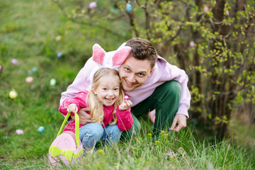 Happy Caucasian family, father and blond cute daughter with bunny ears collecting Easter eggs and putting Chocolate Egg On Easter Egg Hunt In Garden. Easter concept
