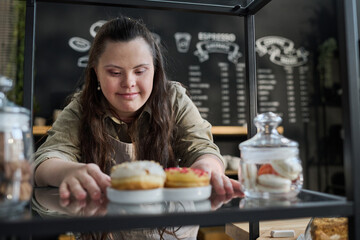 Young smiling waitress with mental disability putting fresh tasty donuts on tray while preparing order of client by counter in front of camera