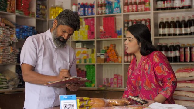 Kirana store merchant noting down orders from customer - successful small businesses  grocery store. Shot of a young Indian woman buying groceries from local grocery shop - monthly ration  inflatio...