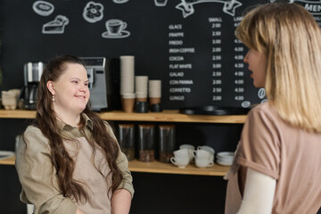 Happy young waitress with Down syndrome and her colleague having chat while standing by workplace...