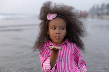 Playful dark-skinned girl with pink hair hoop sends air kiss against background of sea and beach.