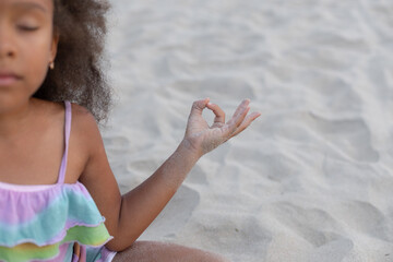 Cropped image of small dark-skinned girl who sits cross-legged on beach on summer day meditates. 