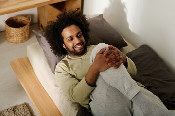 Smiling man in cotton pullover and sweatpants doing stretching exercise for legs after sleep while...