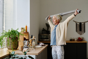 Active senior woman in sportswear holding towel over head between stretched arms while doing physical exercise in the kitchen