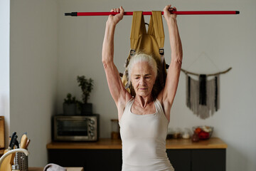 Senior woman with white hair looking in front of herself while holding stick with heavy backpack over head during morning workout