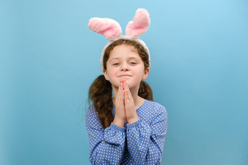 Portrait of pretty little girl child wearing pink easter bunny ears praying with hands together asking for forgiveness smiling confident, posing isolated over blue color background wall in studio