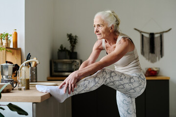 Active senior woman making effort while stretching arms over raised leg on kitchen counter and...