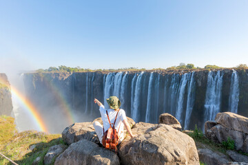 woman sitting on top of a rock points to Victoria Falls - Zimbabwe, travel concept