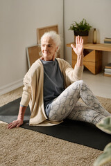 Senior woman in leggins, t-shirt and cardigan keeping left hand raised and right leg bent in knee while sitting on the floor and practicing yoga