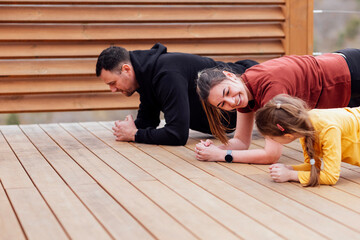 Happy family are stretching outdoors. Attractive mom, handsome dad and cute daughter doing sport exercises. Young couple and their kid are training in park. Smiling man, woman and girl