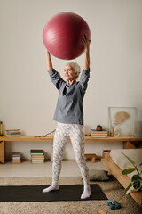 Active retired woman in sportswear holding fitball over her head while standing on the floor of...