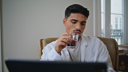 Serious guy enjoying cocktail at home close up. Relaxed man watching laptop