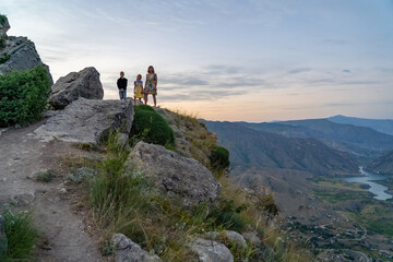Active woman in dress with her son and daughter on cliff in mountains at sunset