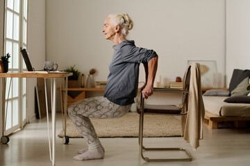 Side view of aged active woman in casualwear doing squats in front of laptop while holding by chair...