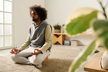 Gordijnen Calm guy in hoodie and sweatpants relaxing on the floor of bedroom while sitting in pose of lotus and meditating in the morning © pressmaster