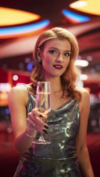 Vertical Screen: Beautiful Caucasian Woman Posing In Prestige Casino Next To Baccarat Table, Wearing Fancy Dress. Female Player Enjoying Glass Of Champagne, Smiling, Looking At Camera in Slow Motion.
