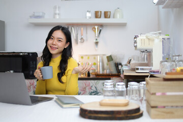 Young Asian woman drinking coffee working with laptop in kitchen at home.