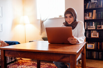 Pretty young middle eastern woman wearing hijab using laptop at home