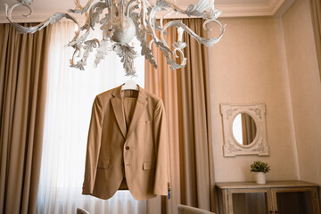 Brown beige jacket hangs on white chandelier. Groom wedding concept. Warm room wedding suite.