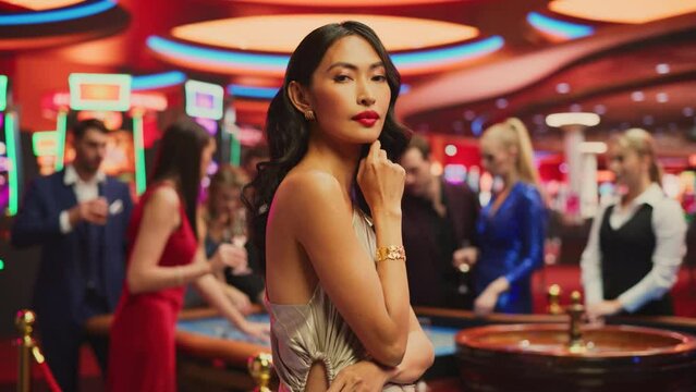 Portrait of a Beautiful Young Asian Woman, Wearing an Elegant Silver Dress, Posing in a Glamorous Atmosphere of a Casino, Surrounded by Active Gamblers Playing Roulette On the Background. Slow Motion