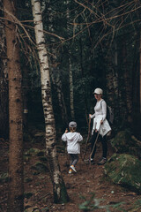An adult girl and her little traveler daughter go into a dense dark forest in the mountains. Trekking poles in hands and backpacks on shoulders