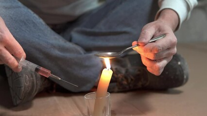 A drug addict cooks heroin on a spoon with the help of a candle fire. Drug addiction and homelessness concept. - Powered by Adobe