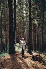 A traveler stands on a rocky mountain path among tall coniferous trees with trekking poles in her hands.