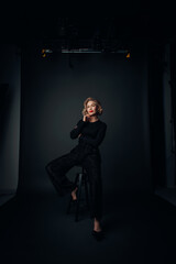 Cool Business Woman dressed i black sitting on a stol in a photo studio - obrazy, fototapety, plakaty