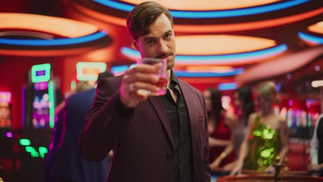 Portrait of a Sophisticated Young Man in an Expensive Suit, Smiling, Looking at Camera and Raising a Whisky Glass in a Luxurious Casino with Gamblers Playing Roulette in Colorful Bokeh Background