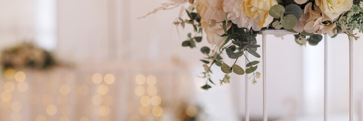A bouquet of white and light yellow roses in a tall white vase with a gold base on a blurred background of a banquet hall. Banner with copy space. - obrazy, fototapety, plakaty
