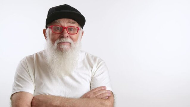 Aged man with grey beard, playfully posing for the camera. He is fashionably dressed in a black cap, red glasses, and a white t-shirt. Arms crossed under the chest. RED 8K RAW.
