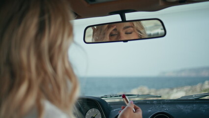 Woman looking car mirror correcting hairstyle close up. Blonde holding lipstick.