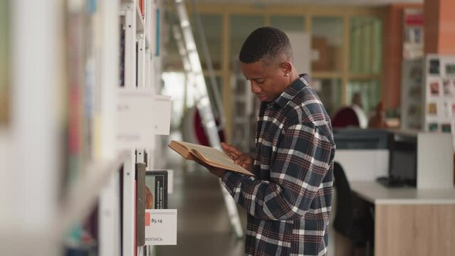 Dark-skinned man peruses book near bookshelf. African American student studies book fluently before picking up for home reading. Guy reads novel