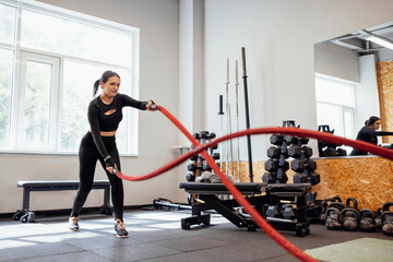 Attractive young slim and fit sportswoman working out in the gym for functional training doing crossfit exercises with battle ropes