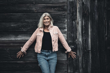 Older woman in her fifties in front of an old wooden wall, dressed in a pink denim jacket, blue jeans and a black T-shirt, in a retro style