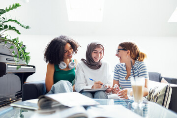Three cute teenage girls do their homework. Caucasian, African American and a Muslim female teens sit at a table with books and notebooks. White wall and plant pots on background. Copy space.
