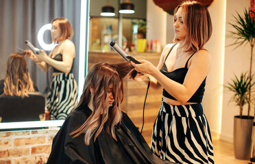 Fototapeta premium Beautiful young woman getting hair done by professional hairstylist in beauty salon