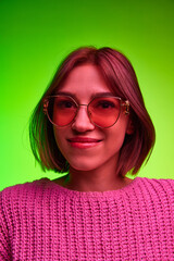 Fototapeta premium Vertical portrait of young beautiful woman in modern, fashion sunglasses looking at camera against green background in pink neon light, filter. Concept of art, beauty, youth, human emotions, style. Ad