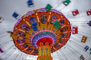Um brinquedo de rotação, grande e colorido, no parque de diversões com poucas pessoas, em um dia nublado. Foto feita de baixo para cima. - obrazy, fototapety, plakaty