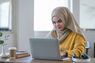 Muslim entrepreneur focused on work at her laptop in a bright office space.