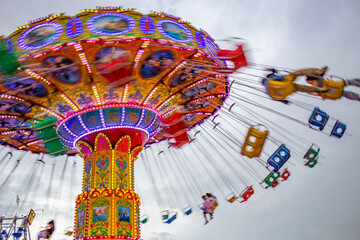 Detalhe de um brinquedo gigante de rotação no parque de diversões com poucas pessoas, em um dia nublado. Foto feita de baixo para cima. - obrazy, fototapety, plakaty