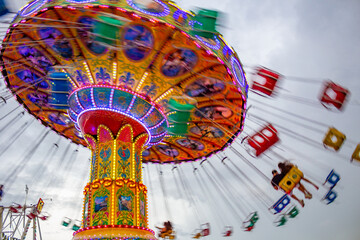 Um brinquedo gigante de rotação no parque de diversões com 2 pessoas em um dos balanços, em um dia nublado. Foto feita de baixo para cima. Imagem feita com baixa velocidade da câmera. - obrazy, fototapety, plakaty