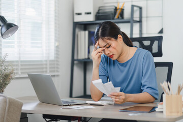 Worried asian woman managing finances on laptop at home office.