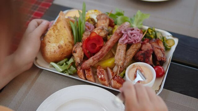 Woman dips piece of grilled sausage into sauce and eats it, view over the shoulder. Large appetizing lunch platter with assorted meats and vegetables in slavic cafe.