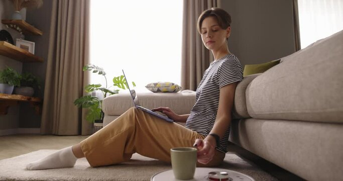 Young woman sitting on floor leaning his back on sofa, working on laptop as robot vacuum cleaner delivered fresh cup of coffee. Modern technology, convenience for freelance lifestyle. Cinematic AD